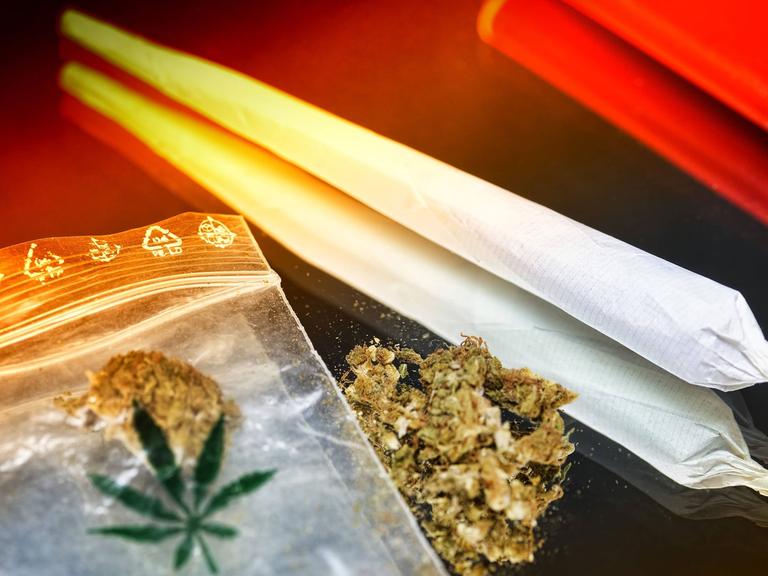 Auf einem Tisch liegt ein Plastiktütchen mit Hanf neben einem Joint und einem roten Feuerzeug.