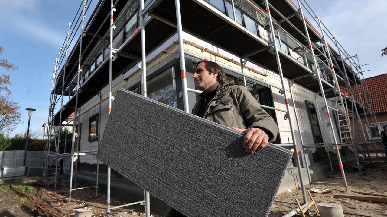 Ein Mann trägt vor dem Rohbau eines mehrstöckigen Hauses eine Wärmedämmplatte unter dem Arm. Ein Mann trägt vor dem Rohbau eines mehrstöckigen Hauses eine Wärmedämmplatte unter dem Arm.