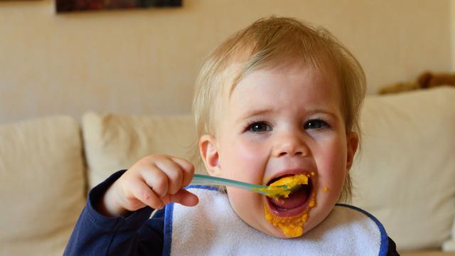 Ein kleines Mädchen löffelt ohne Hilfe allein und mit großem Appetit seinen Möhrenbrei aus einer Plastikschüsseli.