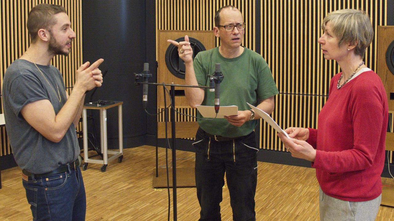 Regisseur Noam Brusilovsky und die Schauspieler Stephan Wolf-Schönburg und Anna Stieblich stehen in einem Hörspiel-Studio und diskutieren. In der Mitte steht ein Mikrofon. Der Raum ist holzgekachelt, im Hintergrund stehen große Lautsprecher. Regisseur Noam Brusilovsky und die Schauspieler Stephan Wolf-Schönburg und Anna Stieblich stehen in einem Hörspiel-Studio und diskutieren. In der Mitte steht ein Mikrofon. Der Raum ist holzgekachelt, im Hintergrund stehen große Lautsprecher.