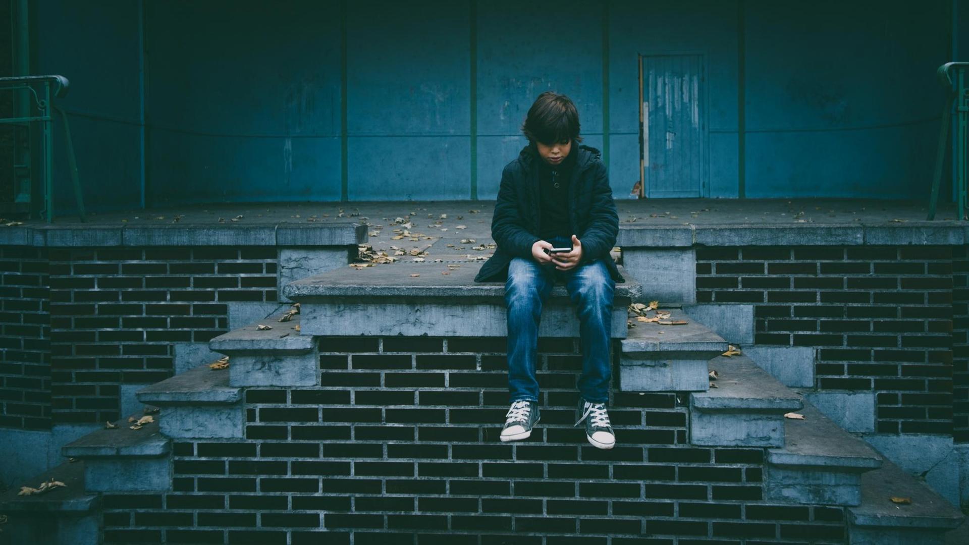 Ein Junge sitzt auf einer Treppe und schaut auf sein Smartphone. Ein Junge sitzt auf einer Treppe und schaut auf sein Smartphone.