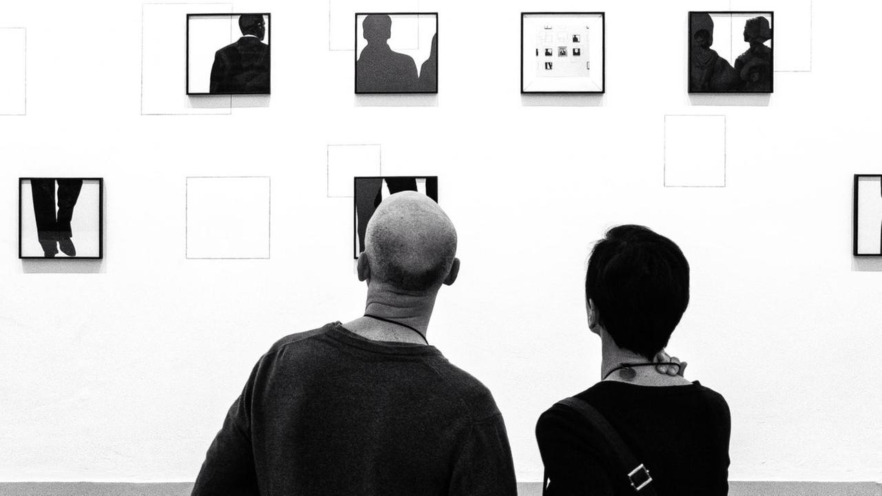 Das schwarz-weiße Bild zeigt zwei Personen, die auf eine Wand mit vielen ungeordneten Bilderrahmen gucken. Das schwarz-weiße Bild zeigt zwei Personen, die auf eine Wand mit vielen ungeordneten Bilderrahmen gucken.