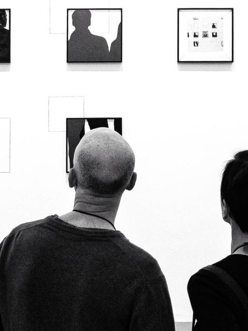 Das schwarz-weiße Bild zeigt zwei Personen, die auf eine Wand mit vielen ungeordneten Bilderrahmen gucken.