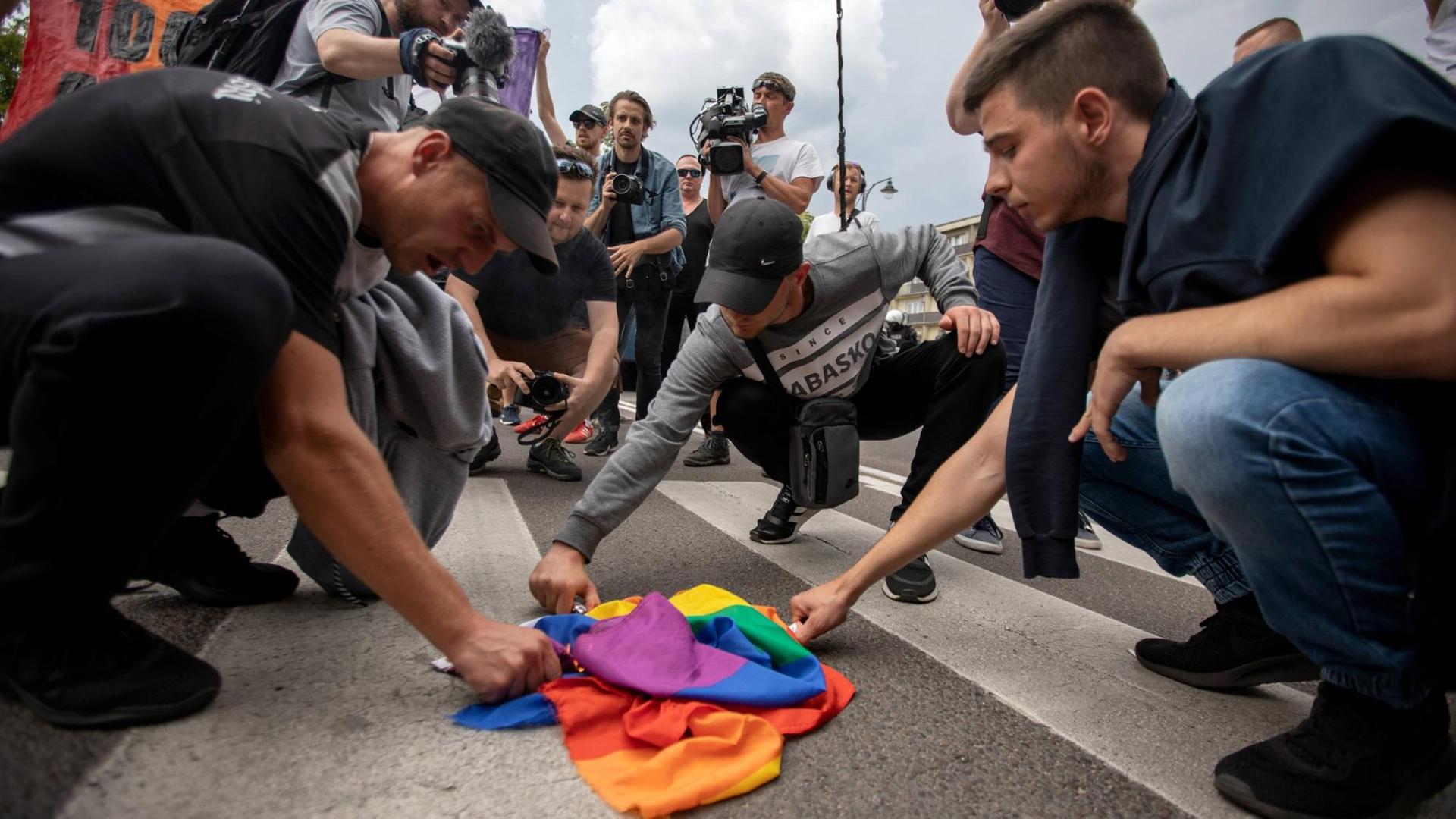 Eine Gruppe junger Männer hält Feuerzeuge an eine Regenbogenflagge, die zusammengeknüllt auf einem Zebrastreifen liegt. Eine Gruppe junger Männer hält Feuerzeuge an eine Regenbogenflagge, die zusammengeknüllt auf einem Zebrastreifen liegt.