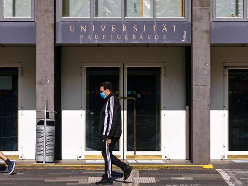 Der fast menschenleere Eingang der Albertus Magnus Universität zu Köln. Nur zwei junge Männer, davon einer mit Mundschutz, laufen an dem Gebäude vorbei.
