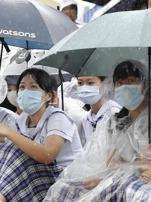 Mit Regenschirmen, Regenumhängen und Munschutz sitzen chinesische Studentinnen bei den Hongkonger Protesten auf der Straße.