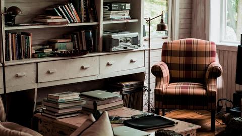 In einem gemütlichen Raum steht ein Sessel neben einem Bücherregal. Im Vordergrund ist ein Schreibtisch zu sehen.