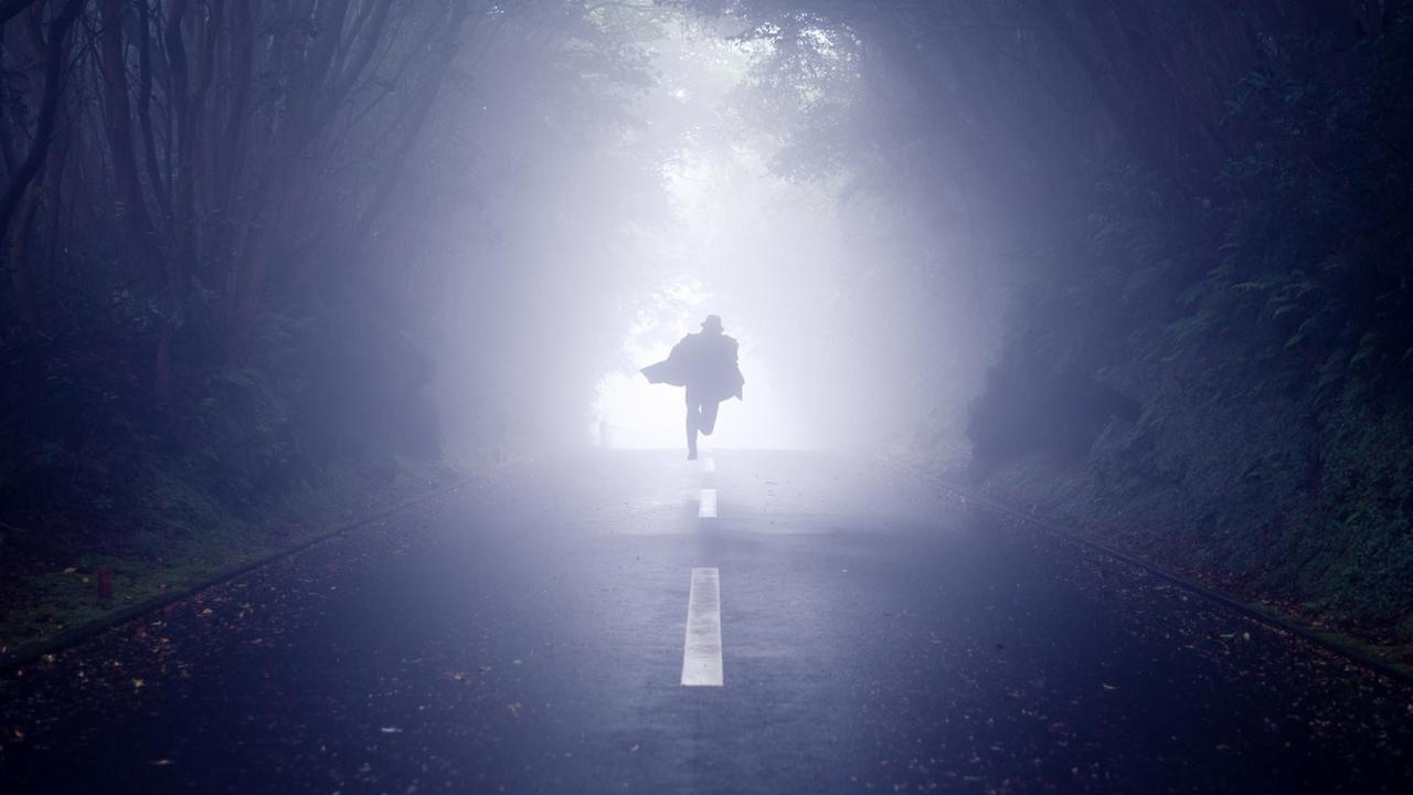 Ein schemenhafter Mann flüchtet auf einer Straße, die durch einen Wald führt