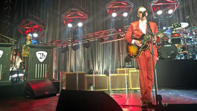 Ein Mann im roten Anzug steht an auf einem Bühne einem Mikrofon, er singt und spielt Gitarre. Im Hintergrund steht ein Schlagzeug und eine ganze Reihe Gitarrenverstärker.