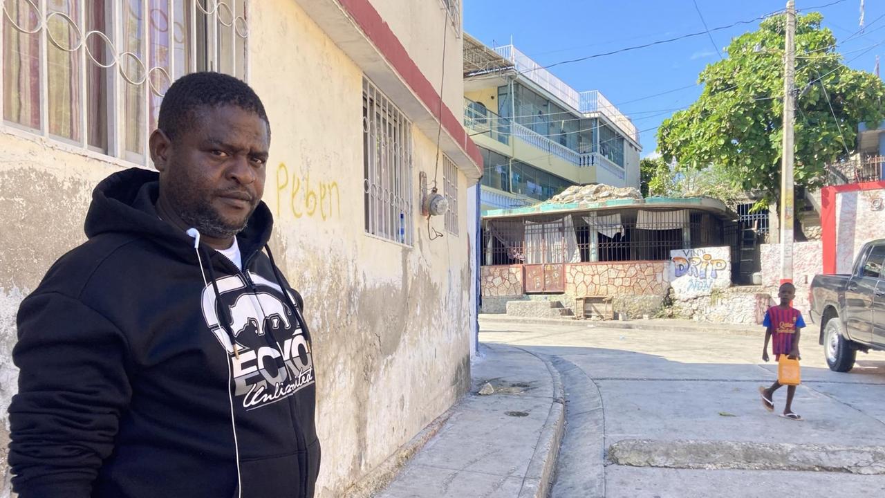 Ein Mann in dunkelblauem Sweater steht neben einem Haus und schaut kritisch. Im Hintergrund geht ein Jungen über die Straße. Ein Mann in dunkelblauem Sweater steht neben einem Haus und schaut kritisch. Im Hintergrund geht ein Jungen über die Straße.