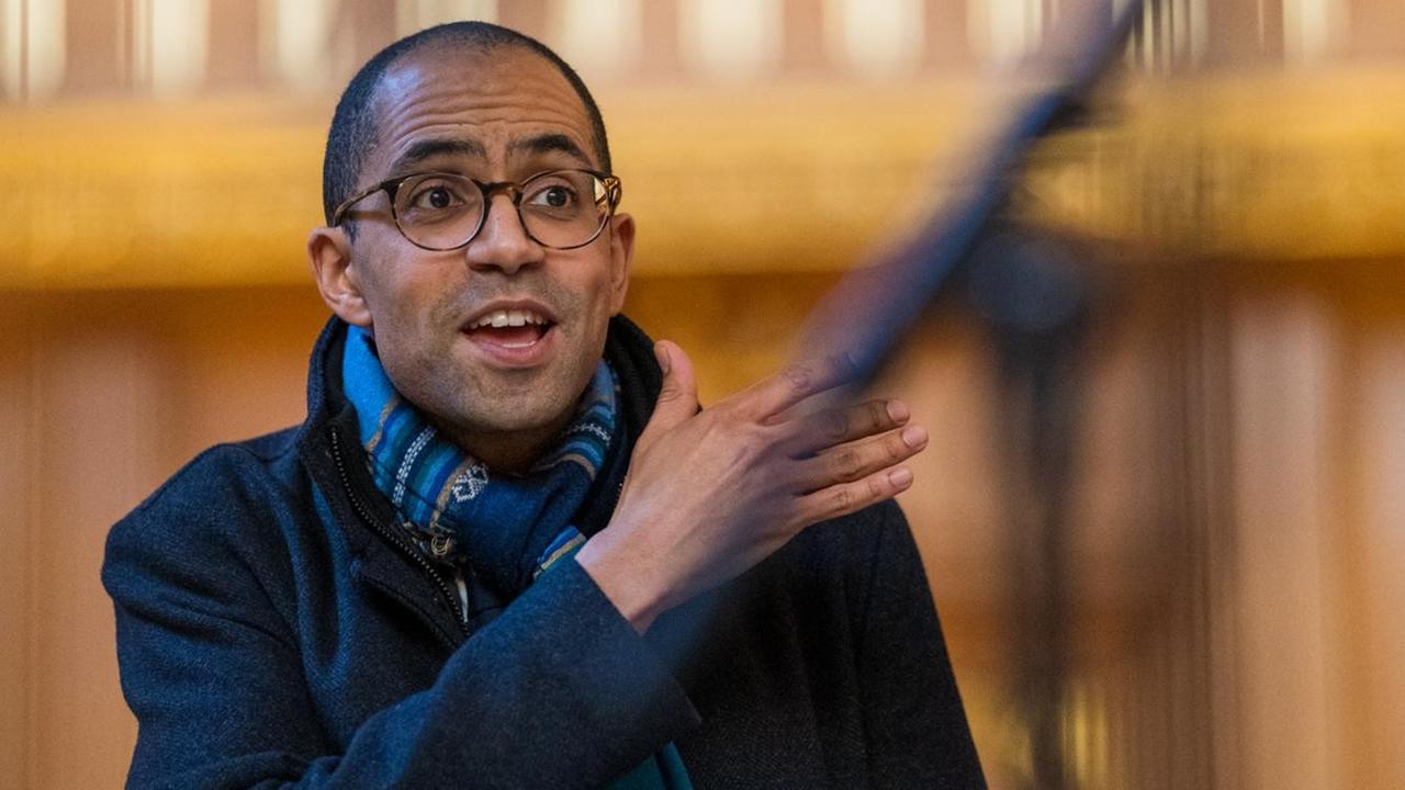 Der schwarze Dirigent dirigiert mit der rechten Hand und schaut auf den Chor, er trägt eine Brille, kurze schwarze Haare und eine blaue Jacke mit einem mittelblauen Schal Der schwarze Dirigent dirigiert mit der rechten Hand und schaut auf den Chor, er trägt eine Brille, kurze schwarze Haare und eine blaue Jacke mit einem mittelblauen Schal