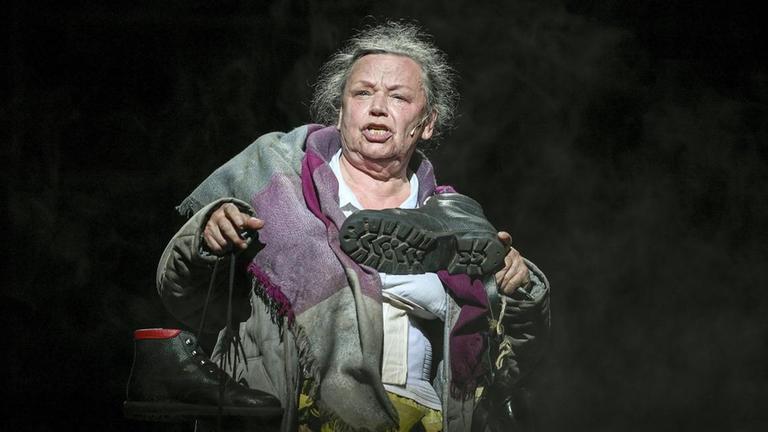 Das Foto ist eine Einzelaufnahme von Ursula Werner in der Rolle der Mutter Courage, in der sie in ihrer linken Hand einen Schuh mit der Schuhsohle in Richtung Kamera hochhält. Das Foto ist eine Einzelaufnahme von Ursula Werner in der Rolle der Mutter Courage, in der sie in ihrer linken Hand einen Schuh mit der Schuhsohle in Richtung Kamera hochhält.
