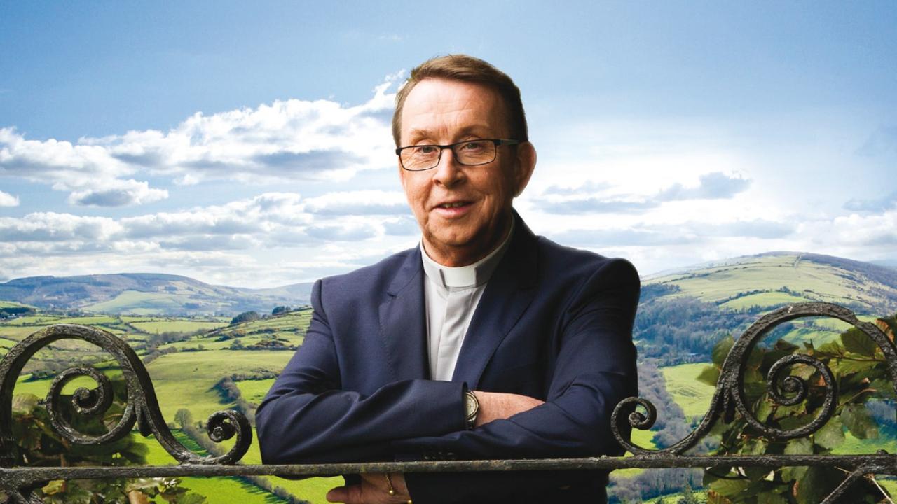 Promofoto von Father Ray Kelly im Rahmen seines Albums "Where I Belong". Es zeigt ihn Priesterkleidung auf einen schmiedeeisernen Zaun gestützt. Im Hintergrund ist eine liebliche Hügellandschaft mit leicht bewölkten blauem Himmel zu sehen.