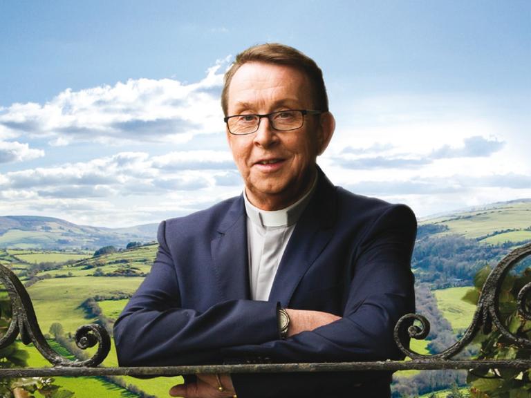 Promofoto von Father Ray Kelly im Rahmen seines Albums "Where I Belong". Es zeigt ihn Priesterkleidung auf einen schmiedeeisernen Zaun gestützt. Im Hintergrund ist eine liebliche Hügellandschaft mit leicht bewölkten blauem Himmel zu sehen.