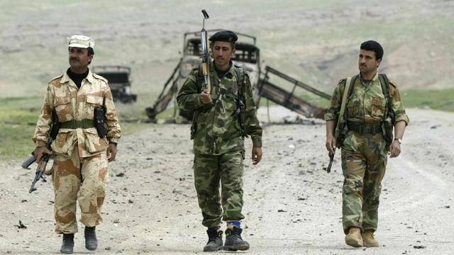 Drei kurdische Peschmerga-Milizen laufen in einer kargen, hügeligen Landschaft nebeneinander her auf den Betrachter zu.