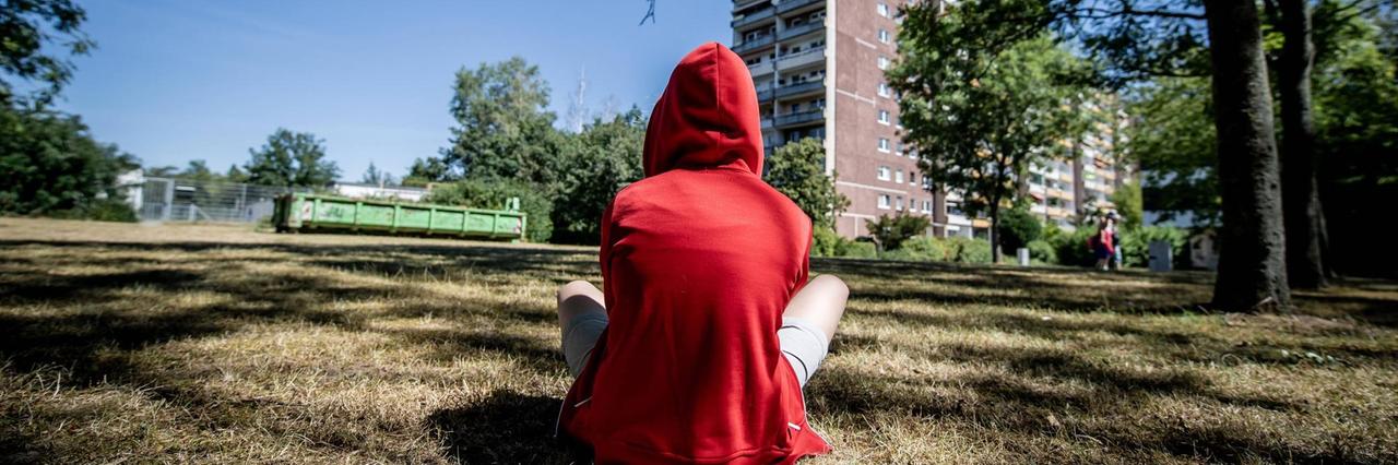 Ein jugendliches Kind sitzt in einem roten Kapuzenpullover mit dem Rücken zum Betrachter auf einer Wiese. Im Hintergrund ist ein Plattenbau zu sehen. Ein jugendliches Kind sitzt in einem roten Kapuzenpullover mit dem Rücken zum Betrachter auf einer Wiese. Im Hintergrund ist ein Plattenbau zu sehen.