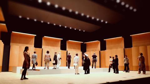 Die Mitglieder des Ensembles stehen wild verteilt auf einem modernen Bühnenraum