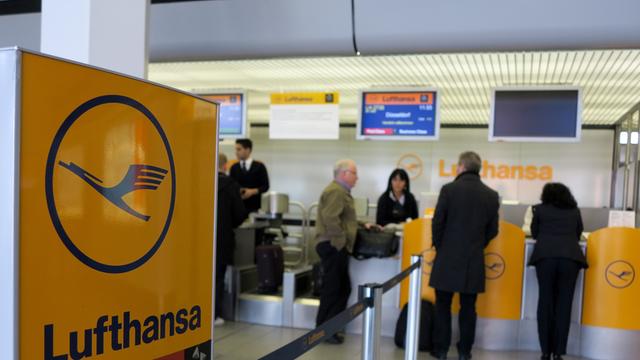 Check-in-Schalter der Lufthansa, im Vordergrund ein Lufthansa-Schild, im Hintergrund der Schalter mit Mitarbeitern und Passagieren.