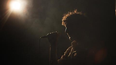 Ein Mann steht auf dunklen Bühne und singt in ein Mikrofon.