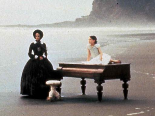 Eine Frau steht an einem Strand, neben ihr liegt ein Mädchen auf einem Piano.