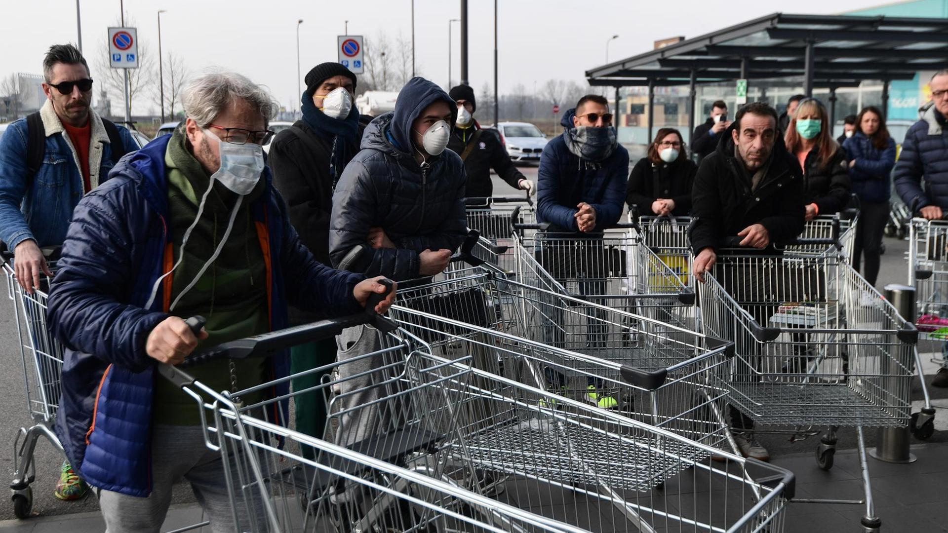 Eine Gruppe Menschen mit Atemschutzmasken steht mit leeren Einkaufswagen auf einem Parkplatz. Eine Gruppe Menschen mit Atemschutzmasken steht mit leeren Einkaufswagen auf einem Parkplatz.