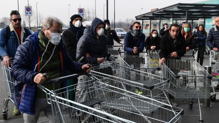 Eine Gruppe Menschen mit Atemschutzmasken steht mit leeren Einkaufswagen auf einem Parkplatz.