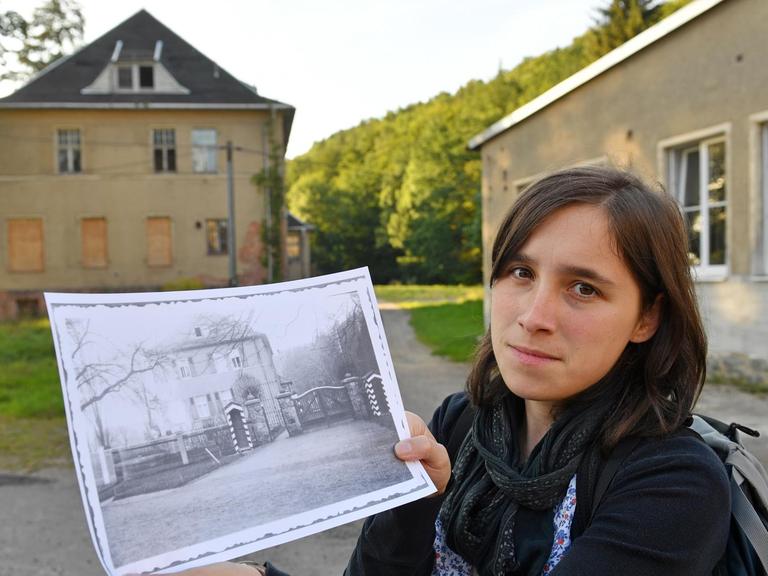 Vor der mittlerweile verfallenen Kommandantenvilla des ehemaligen KZ Sachsenburg in Frankenberg (Sachsen) hält Anna Schüller ein historisches Foto der Villa in ihren Händen.