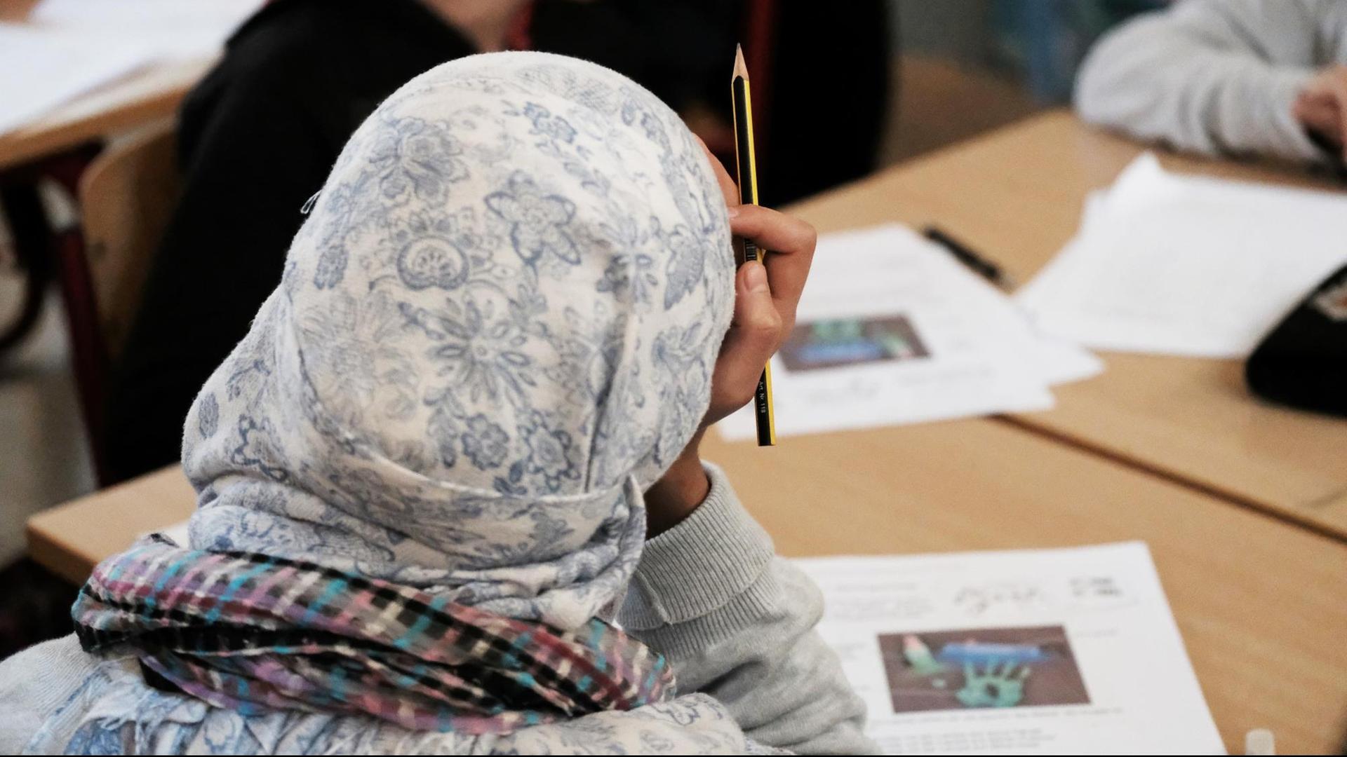 Ein Mädchen mit Kopftuch sitzt über ein Arbeitsblatt gebeugt an einem Pult im Klassenraum. im Hintergrund weitere Schüler.