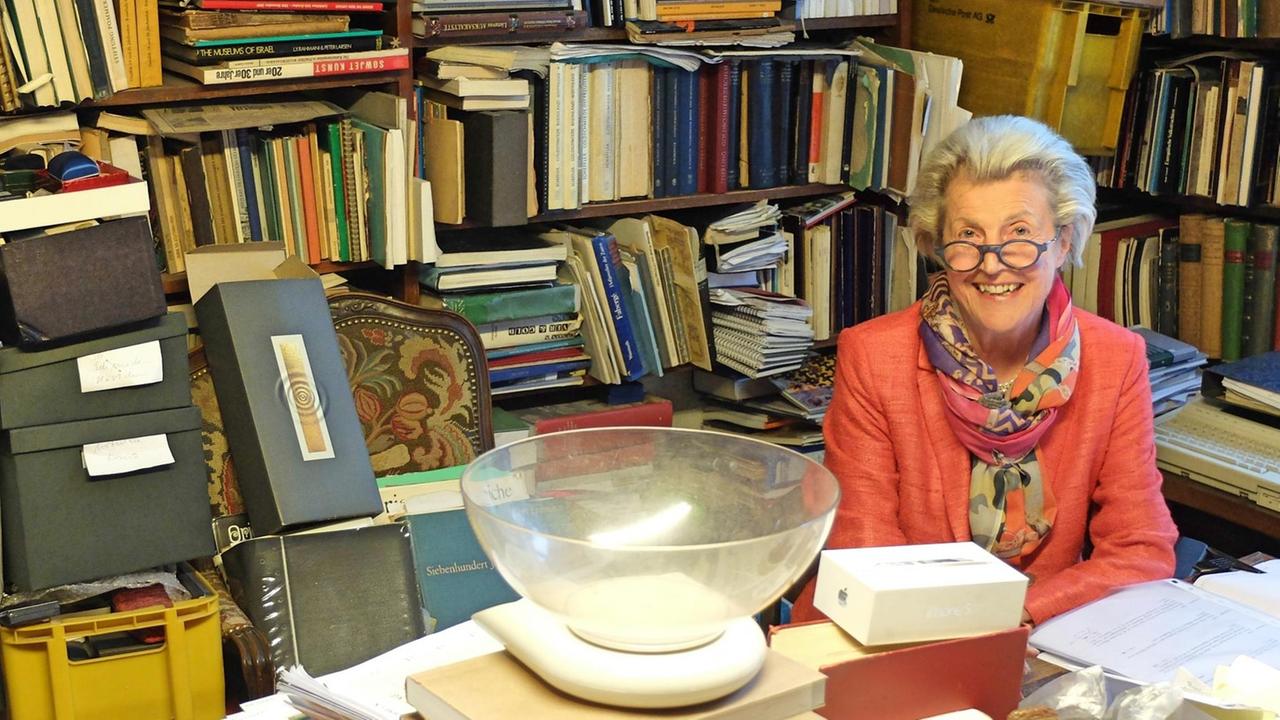 Eine ältere Dame sitzt in einem mit Büchern vollgestellten Büro. 