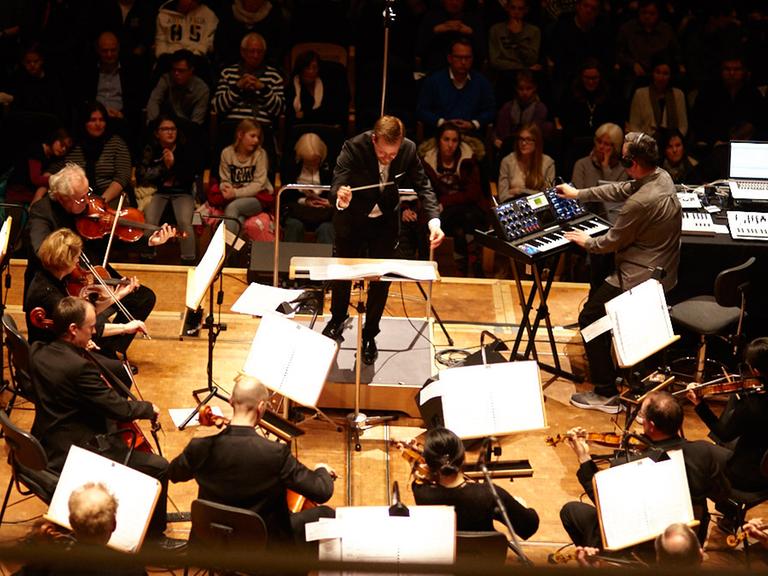 Mark Romboy und die Dortmunder Philharmoniker präsentieren ihre Version von Debussy. Man sieht Streicher und Dirigent vor Publikum sowie Mark Romboy am Synthesizer.