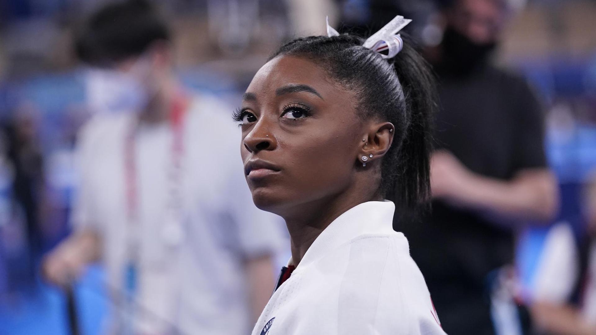 Eine schwarze Athletin im weißem Oberteil blickt ernst an der Kamera vorbei. Es ist die Turnerin Simone Biles. Eine schwarze Athletin im weißem Oberteil blickt ernst an der Kamera vorbei. Es ist die Turnerin Simone Biles.