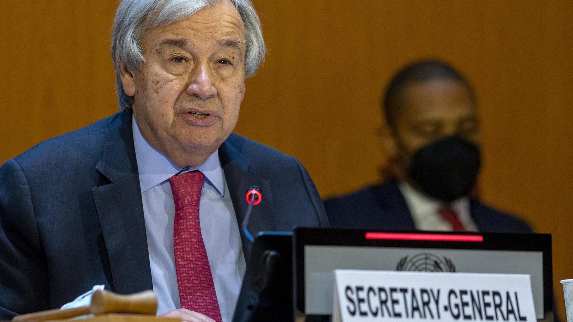 Guterres spricht in ein Mikrofon. Vor seinem Platz steht ein Schild mit der Aufschrift "Secretary-General". Dahinter unscharf ein Mann mit schwarzer Gesichtsmaske.