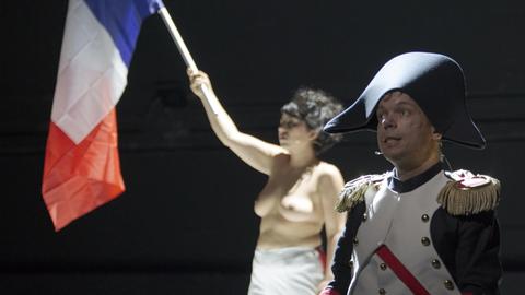 Ein Darsteller im Napoleonskostüm steht auf der Bühne. Im Hintergrund sieht man eine barbusige Frau, die die Frankreichflagge schwenkt.