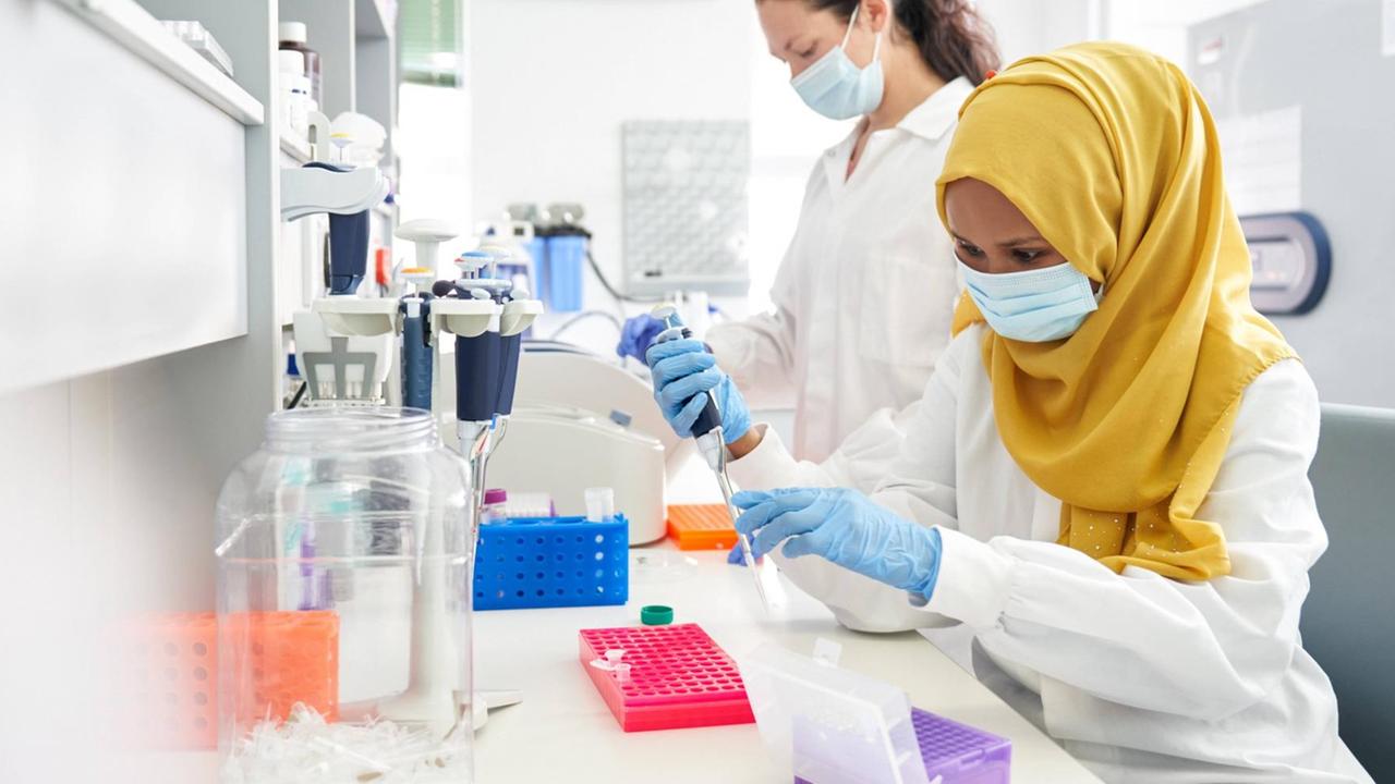 Zwei Wissenschaftlerinnen mit Maske, eine mit Hijab, arbeiten in einem Labor Zwei Wissenschaftlerinnen mit Maske, eine mit Hijab, arbeiten in einem Labor