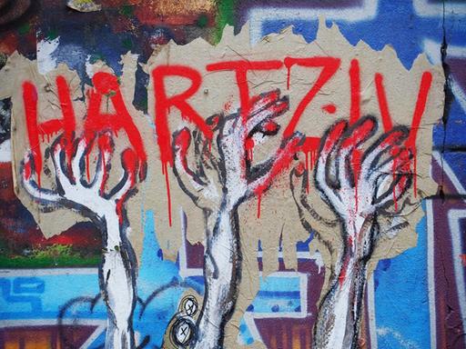 Ein unbekannter Street-Art-Künstler hat auf einer Mauer an einem besetzten Haus in Berlin im Bezirk Mitte dieses Bild geschaffen, das seine Meinung zu den Hartz IV Gesetzen drastisch wiedergibt: Verzwifelte HÄnde greifen nach dem roten Schriftzug Hartz IV, im Hintergrund sind Grabkreuze zu sehen.