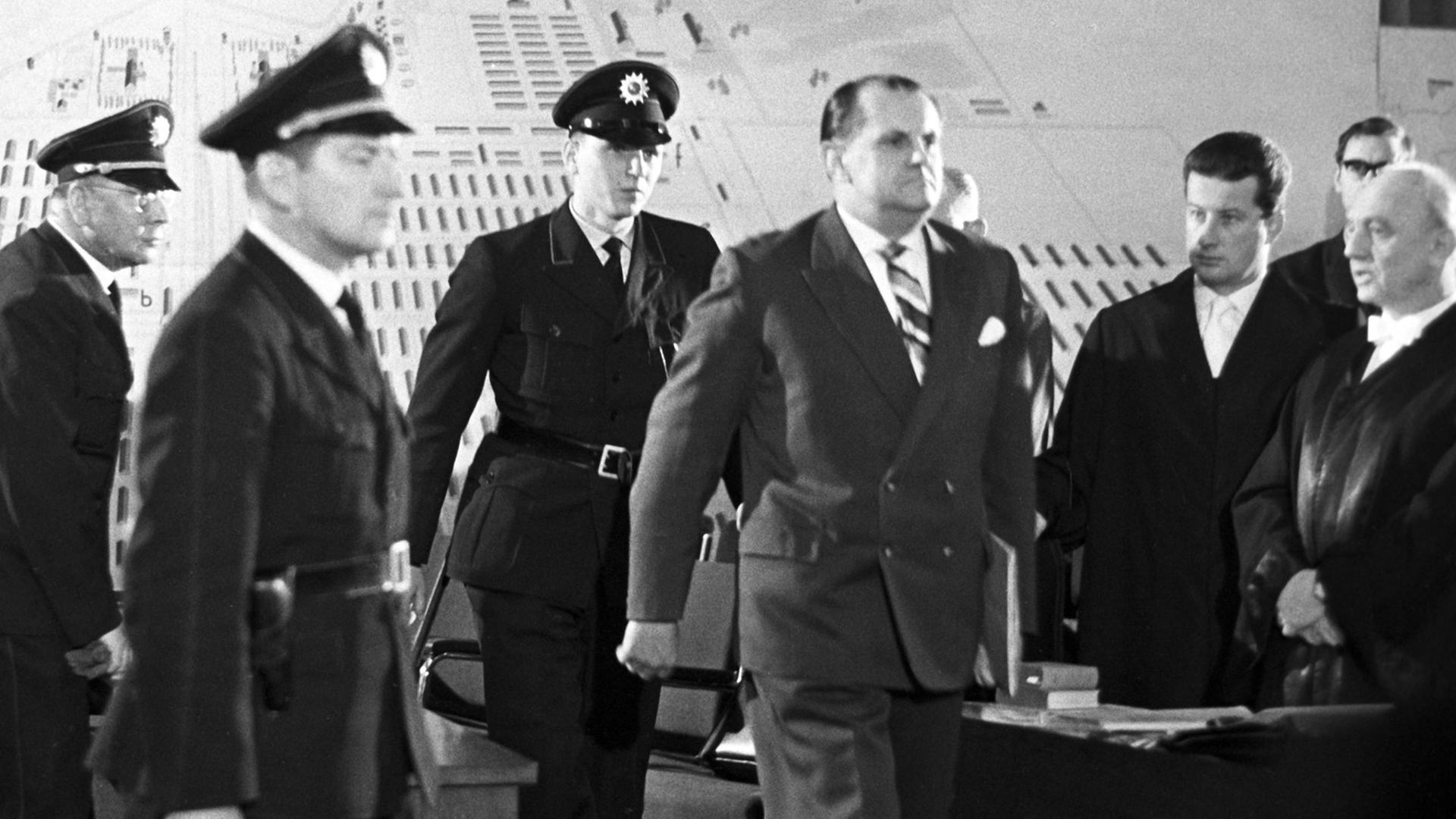 Der Angeklagte Oswald Kaduk wird von drei Polizisten in einen Gerichtssaal geführt. Der Angeklagte Oswald Kaduk wird von drei Polizisten in einen Gerichtssaal geführt.