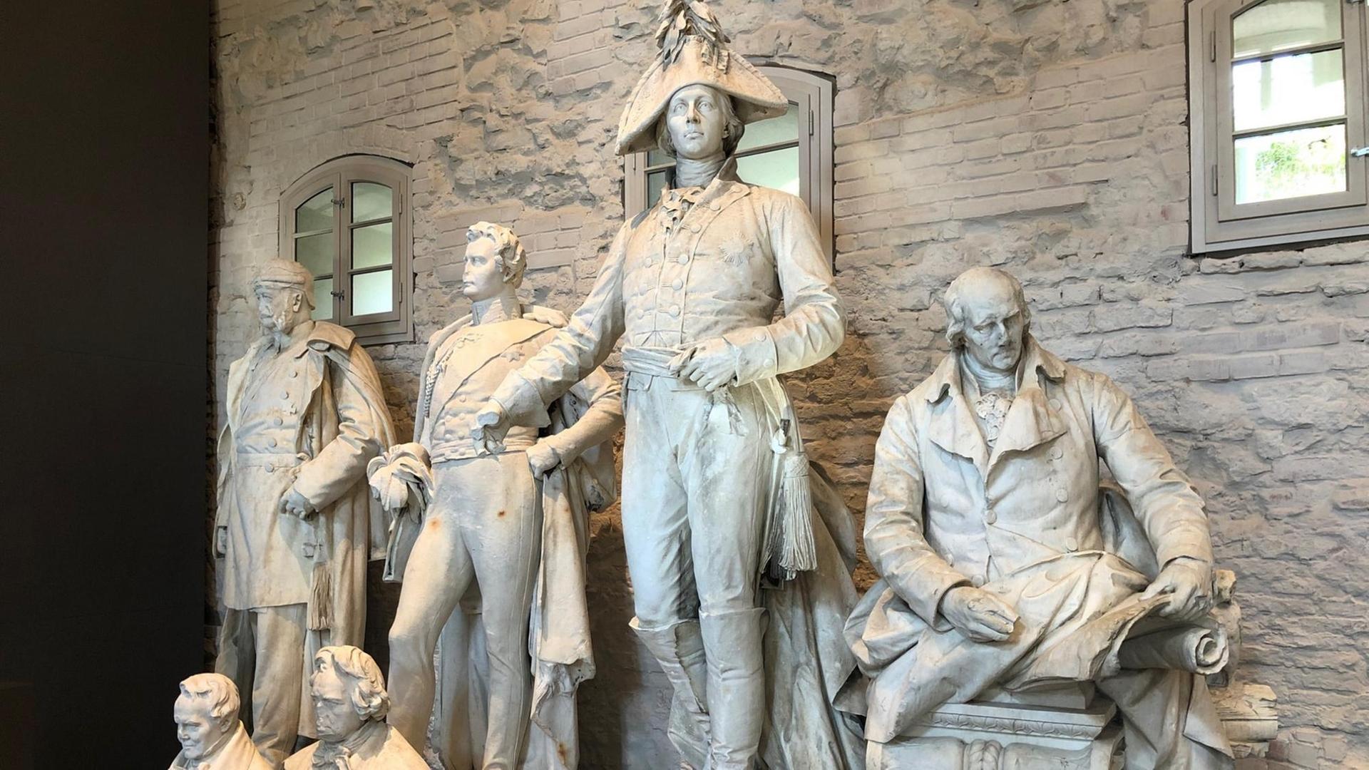 Mehrere steinerne Statuen von Königen und Reformern stehen aufgereiht nebeneinander und bilden ein Ensemble. Mehrere steinerne Statuen von Königen und Reformern stehen aufgereiht nebeneinander und bilden ein Ensemble.