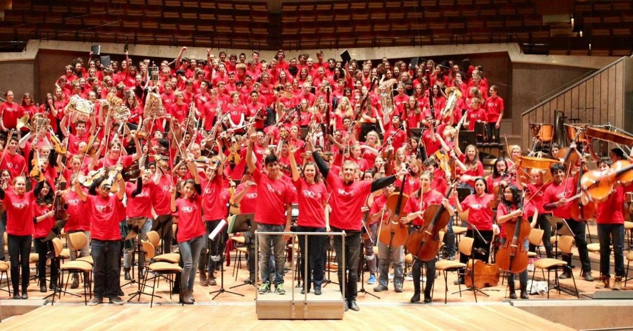 Ein großes Jugendorchester und ein Chor stehen mit roten Shirts auf der Bühne und heben die Arme, zum Teil mit ihren Instrumenten.
