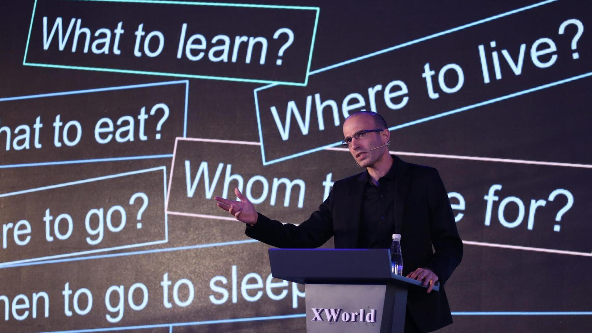 Der Historiker und Bestseller-Autor Yuval Noah Harari auf einer Bühne, im Hintergrund weiße Sätze auf schwarzem Grund mit Fragen wie "Where to live", "What to learn" und Ähnlichem. Der Historiker und Bestseller-Autor Yuval Noah Harari auf einer Bühne, im Hintergrund weiße Sätze auf schwarzem Grund mit Fragen wie "Where to live", "What to learn" und Ähnlichem.