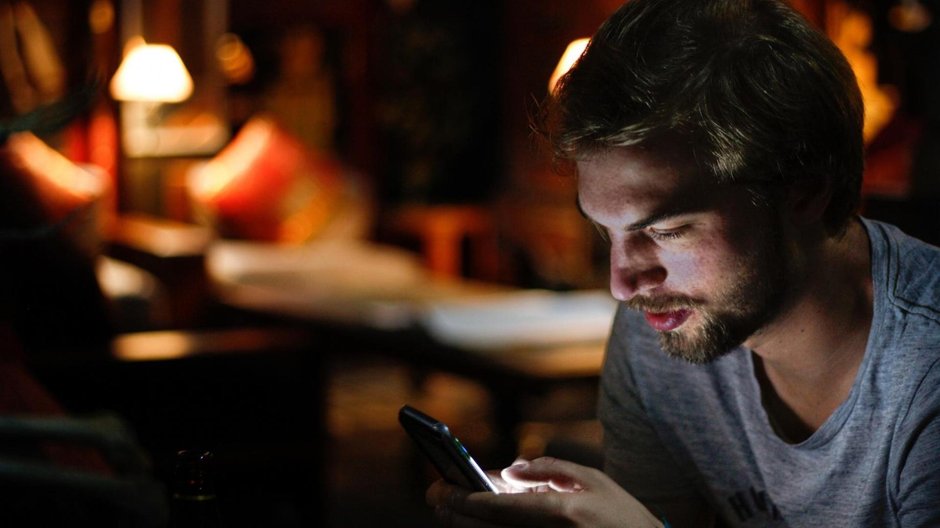 Während ein junger Mann auf sein Smartphone schaut, beleuchtet das Display sein Gesicht. Während ein junger Mann auf sein Smartphone schaut, beleuchtet das Display sein Gesicht.