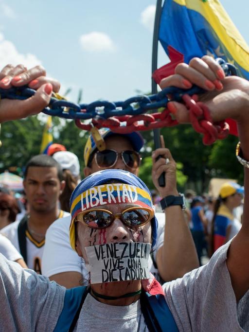 Ein Gegner von Präsident Maduro steht vor weiteren Demonstranten. Er hält eine Kette in seinen Händen; Zudem trägt er ein Stirnband mit der Aufschrift "Libertad".