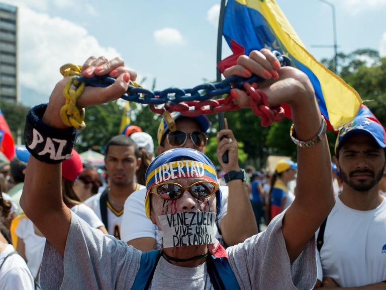 Ein Gegner von Präsident Maduro steht vor weiteren Demonstranten. Er hält eine Kette in seinen Händen; Zudem trägt er ein Stirnband mit der Aufschrift "Libertad".