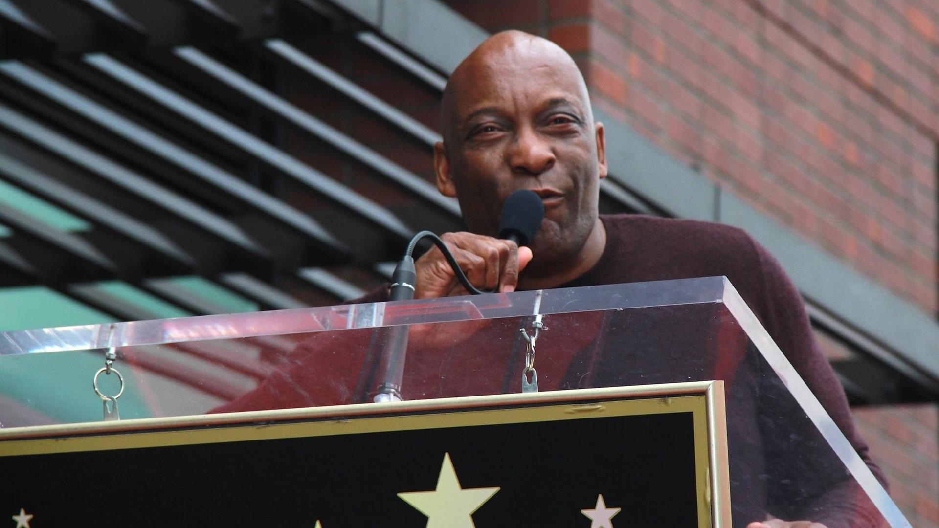 John Singleton steht an einem Redepult mit Mikrofon. John Singleton steht an einem Redepult mit Mikrofon.
