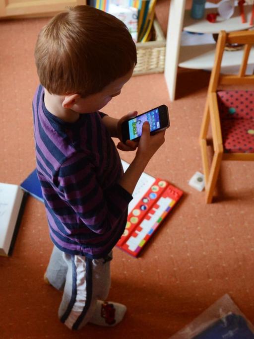 Ein fünfjähriger Junge spielt in seinem unaufgeräumten Kinderzimmer auf einem iPhone ein Computerspiel.