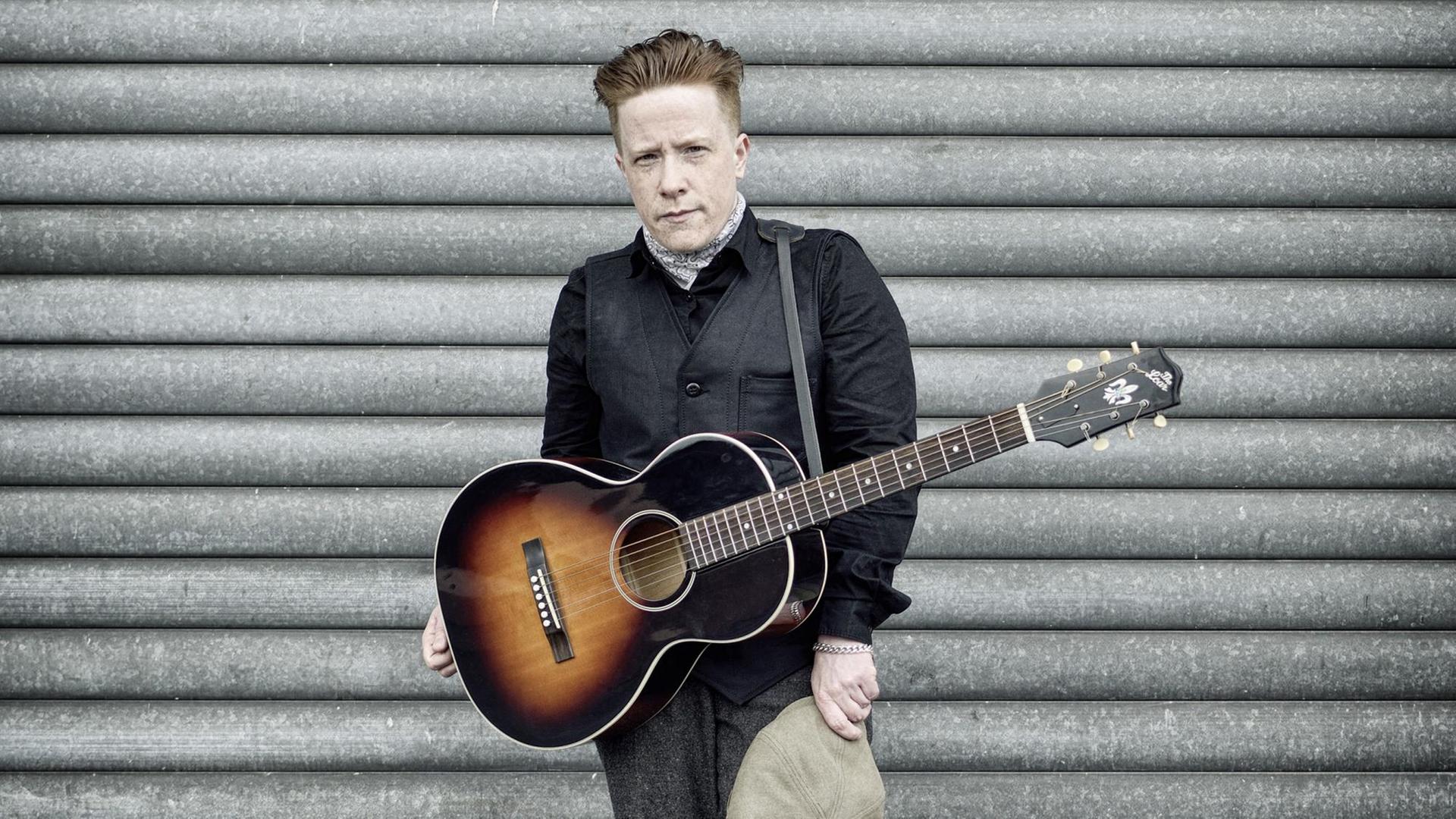 Remco steht mit einer umgehängten Gitarre vor einer Wand. Remco steht mit einer umgehängten Gitarre vor einer Wand.