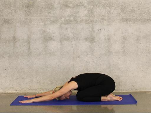 Eine sportliche Frau macht eine Yoga-Übung auf einer blauen Matte vor einer Sichtbeton-Wand.