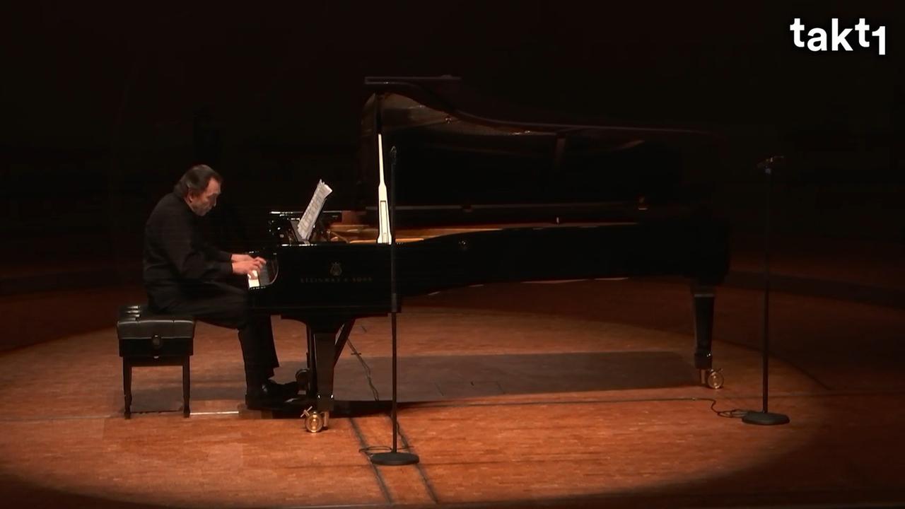 Ein Pianist sitzt an einem langen Konzertflügel. Er ist ganz schwarz gekleidet, der Flügel ist auch schwarz. Er blickt konzentriert auf die Noten vor ihm. Ein Pianist sitzt an einem langen Konzertflügel. Er ist ganz schwarz gekleidet, der Flügel ist auch schwarz. Er blickt konzentriert auf die Noten vor ihm.