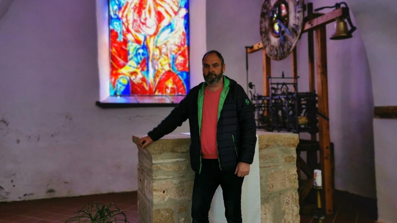 Ein Mann stützt sich auf ein Taufbecken, dahinter ein farbiges Kirchenfenster.