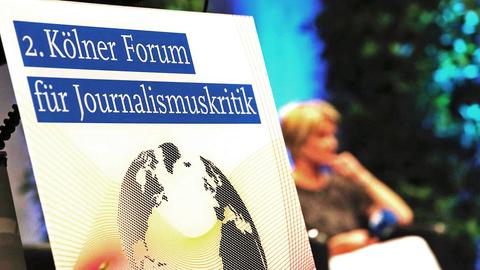 Ein Plakat für das 2. Kölner Forum für Journalismuskritik, im Hintergrund verschwommen ein Gast auf dem Podium