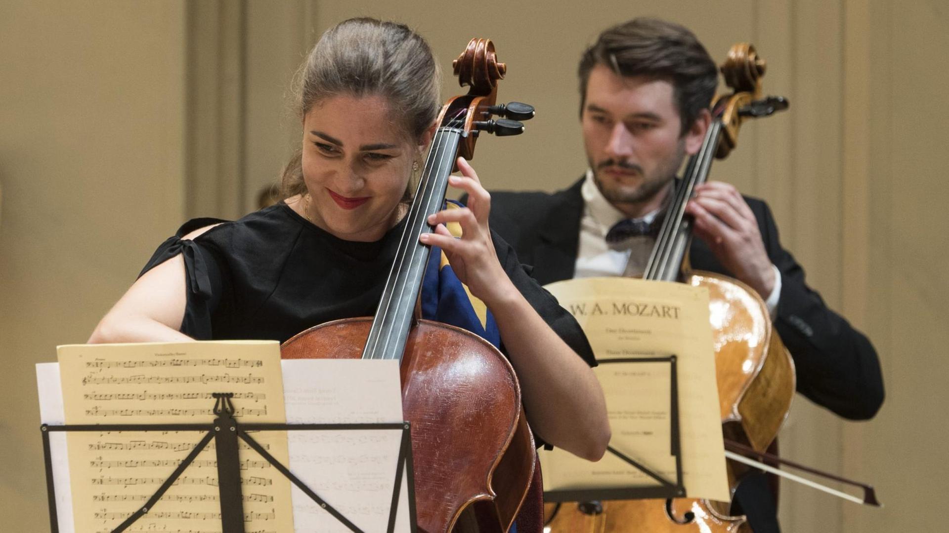 Eine Cellist sitzt vor einem weiteren Cellisten, die beide aus Mozart-Noten spielen. Eine Cellist sitzt vor einem weiteren Cellisten, die beide aus Mozart-Noten spielen.